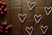 Christmas candy background pattern with red and white striped candy canes arranged as heart shapes on brown tiles with red Christmas baubles, overhead view